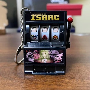 May include: Black miniature slot machine keychain featuring the text "THE BINDING OF ISAAC" in yellow. The slot machine has three reels with various game symbols and a pull lever with a red ball. The bottom of the machine has several cartoon character graphics.