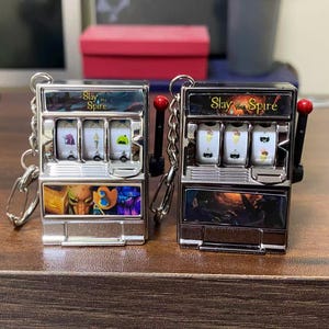 Puede incluir: Dos llaveros plateados con forma de máquina tragamonedas. Cada llavero tiene tres rodillos giratorios con varios símbolos y el texto "Slay the Spire" en la parte superior. Las máquinas tragamonedas tienen un mango y un llavero.