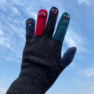 Peut inclure: Un gant en tricot gris foncé avec des visages de monstres colorés sur le bout des doigts. Les bouts des doigts sont bleus, rouges, noirs et sarcelle, chacun avec des yeux et des bouches brodés. Le gant est tenu contre un ciel bleu.