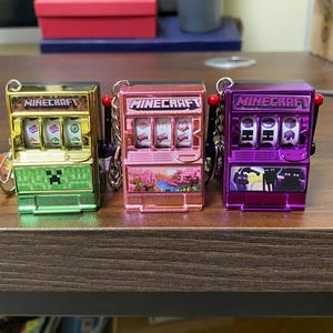 May include: Three miniature slot machine keyrings in green, pink, and purple. Each has the word "Minecraft" on top. The green one has a Creeper face, the pink one has a landscape, and the purple one has a silhouette.