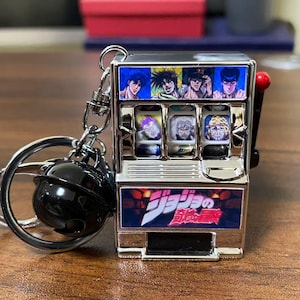 Puede incluir: Un llavero de máquina tragamonedas en miniatura de color plateado con retratos de personajes y texto japonés. El llavero incluye un dije de bola negra y un llavero plateado. La máquina tragamonedas tiene un mango rojo.