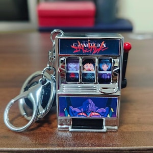 Peut inclure: Un porte-clés argenté avec un design de machine à sous miniature. Le haut de la machine porte le mot "EVANGELION" en rouge et blanc. Les fenêtres de la machine à sous affichent des images de personnages d'anime. Un levier rouge est sur le côté.