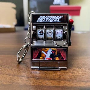 Peut inclure: Un porte-clés en forme de machine à sous argentée avec un levier rouge. La machine à sous comporte trois rouleaux avec des illustrations de personnages et du texte japonais en haut. Le bas de la machine présente une illustration de personnage derrière des rideaux rouges.