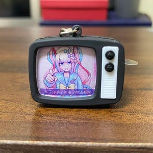 May include: A black miniature television keychain with a cartoon image on the screen. The screen displays a colorful anime character with long pink hair. The text on the bottom of the screen is in a foreign language.