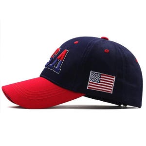 May include: A navy blue baseball cap with a red bill. The word "USA" is embroidered in red and blue on the front. An American flag patch is on the side. The cap has a curved bill and a button on top.
