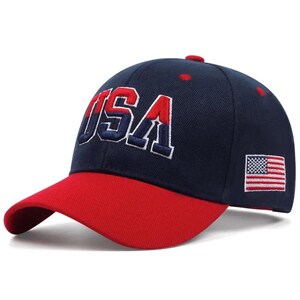 May include: Navy blue baseball cap with a red brim. The cap features the embroidered word "USA" in white and red, along with a small American flag on the side. A classic, patriotic accessory.