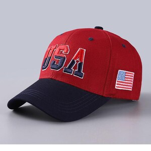 May include: Red baseball cap with a navy blue brim. The cap features the embroidered letters "USA" in red and navy blue, and a small American flag patch on the side. A classic, sporty accessory.