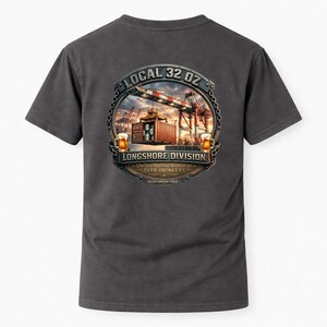 May include: Dark gray t-shirt featuring a detailed graphic. The graphic includes the text "LOCAL 32 OZ" and "LONGSHORE DIVISION" within a circular emblem. The emblem depicts a container being lifted by a crane, with two mugs of beer on either side.