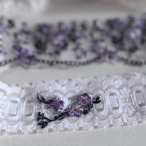 K&ouml;nnte beinhalten: Wei&szlig;er Spitzenstrumpfhalter mit gestickten lila Blumen und gr&uuml;nen Stielen. Der Strumpfhalter hat einen gewellten Rand und ein zartes Blumendesign. Ein zweiter Strumpfhalter ist im Hintergrund zu sehen.