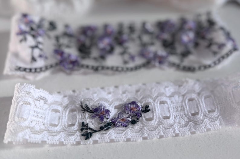 K&ouml;nnte beinhalten: Wei&szlig;er Spitzenstrumpfhalter mit gestickten lila Blumen und gr&uuml;nen Stielen. Der Strumpfhalter hat einen gewellten Rand und ein zartes Blumendesign. Ein zweiter Strumpfhalter ist im Hintergrund zu sehen.