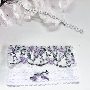 K&ouml;nnte beinhalten: Zwei Teile wei&szlig;er Spitzentrimm mit floraler Stickerei. Das obere St&uuml;ck hat einen gewellten Rand mit lila Blumen und gr&uuml;nen Bl&auml;ttern. Das untere St&uuml;ck ist ein wei&szlig;es Spitzenband mit floralem Design. Zarte wei&szlig;e Spitzentrimm im Hintergrund.