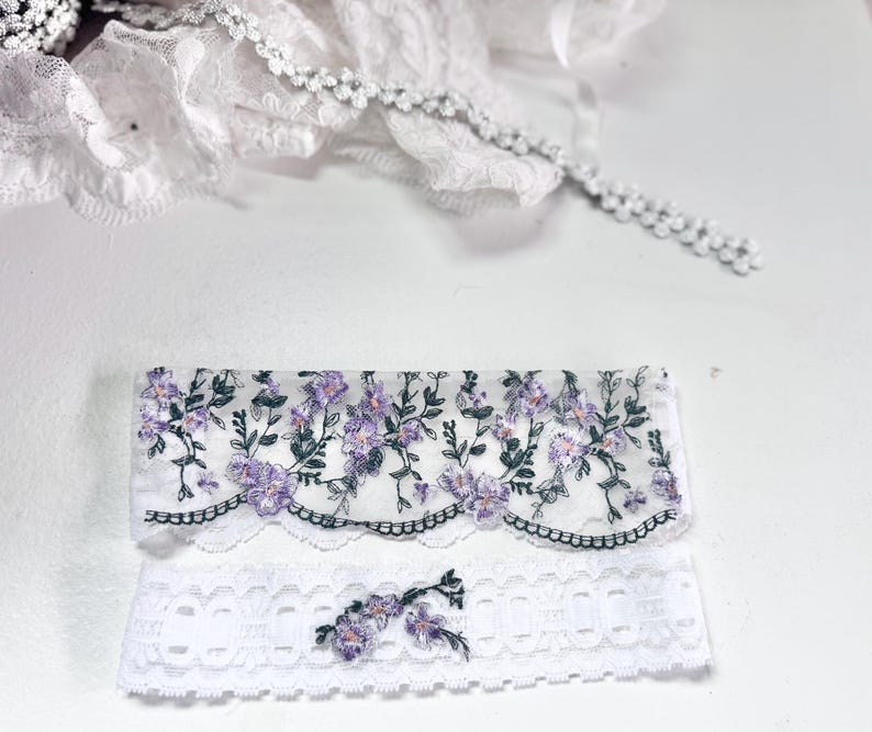 K&ouml;nnte beinhalten: Zwei Teile wei&szlig;er Spitzentrimm mit floraler Stickerei. Das obere St&uuml;ck hat einen gewellten Rand mit lila Blumen und gr&uuml;nen Bl&auml;ttern. Das untere St&uuml;ck ist ein wei&szlig;es Spitzenband mit floralem Design. Zarte wei&szlig;e Spitzentrimm im Hintergrund.