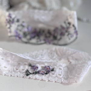 K&ouml;nnte beinhalten: Zwei wei&szlig;e Spitzenstrumpfb&auml;nder mit floraler Stickerei. Die Strumpfb&auml;nder weisen zarte Spitzendetails auf und sind mit kleinen lila und gr&uuml;nen gestickten Blumen verziert. Das Design ist elegant und f&uuml;r besondere Anl&auml;sse geeignet.