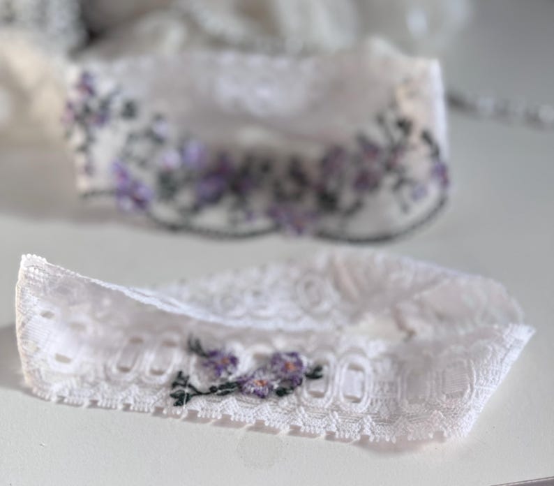 K&ouml;nnte beinhalten: Zwei wei&szlig;e Spitzenstrumpfb&auml;nder mit floraler Stickerei. Die Strumpfb&auml;nder weisen zarte Spitzendetails auf und sind mit kleinen lila und gr&uuml;nen gestickten Blumen verziert. Das Design ist elegant und f&uuml;r besondere Anl&auml;sse geeignet.