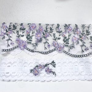 K&ouml;nnte beinhalten: Zwei St&uuml;cke wei&szlig;er Spitzenstoff. Das obere St&uuml;ck ist ein durchsichtiger Stoff mit gestickten lila Blumen und gr&uuml;nen Bl&auml;ttern, der mit schwarzen, gewellten Bord&uuml;ren versehen ist. Das untere St&uuml;ck ist wei&szlig; mit einem floralen Design und gewellten Kanten.