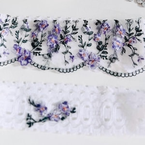 K&ouml;nnte beinhalten: Zwei St&uuml;cke wei&szlig;er Spitzenstoff. Das obere St&uuml;ck hat einen gewellten Rand und ist mit lila und rosa Blumen und gr&uuml;nen Bl&auml;ttern bestickt. Das untere St&uuml;ck ist eine schlichte wei&szlig;e Spitze mit floraler Stickerei.
