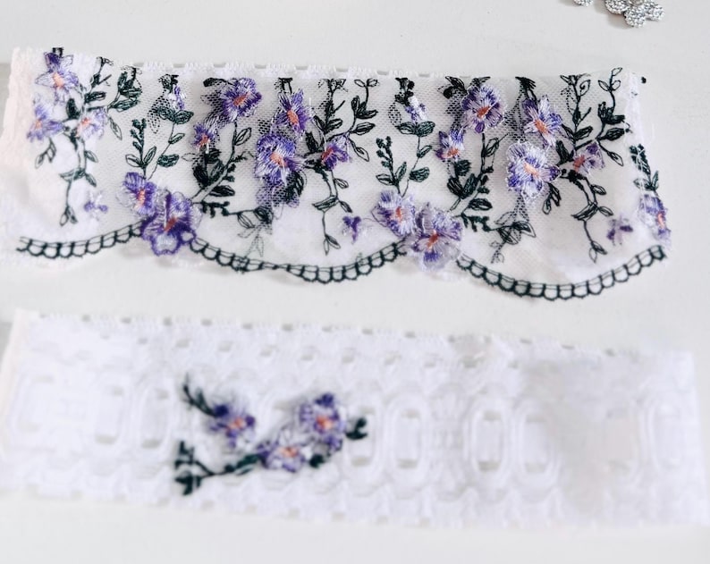 K&ouml;nnte beinhalten: Zwei St&uuml;cke wei&szlig;er Spitzenstoff. Das obere St&uuml;ck hat einen gewellten Rand und ist mit lila und rosa Blumen und gr&uuml;nen Bl&auml;ttern bestickt. Das untere St&uuml;ck ist eine schlichte wei&szlig;e Spitze mit floraler Stickerei.
