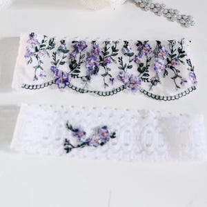 K&ouml;nnte beinhalten: Zwei wei&szlig;e Spitzenstrumpfb&auml;nder mit floraler Stickerei. Das obere Strumpfband hat einen gewellten Rand und ist mit lila Blumen und gr&uuml;nen Bl&auml;ttern verziert. Das untere Strumpfband hat ein kleineres Blumenmuster. Im Hintergrund ist ein silbernes Perlenaccessoire zu sehen.
