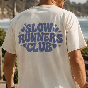 以下が含まれることがあります： クリーム色とライトピンクの半袖Tシャツ2枚。背中には波打つような青いフォントで「SLOW RUNNERS CLUB」の文字がプリントされ、小さな青いハートと星のアイコンが添えられています。どちらも黒いショートパンツと組み合わされています。