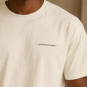 以下が含まれることがあります： 「professional yapper」の文字が小さく刺繍されたアイボリー色のTシャツ。クルーネックと半袖です。全体的なスタイルはカジュアルでリラックスしています。
