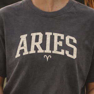 May include: Dark grey t-shirt with the word "ARIES" in white block letters and the Aries zodiac symbol. The person is holding a lacrosse stick with a yellow ball in the net. The shirt is from Wholesome Tees Shop.