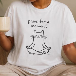 以下が含まれることがあります： 蓮華座で瞑想する猫の黒い線画が描かれた白い半袖Tシャツ。猫の上にはシンプルな黒いフォントで「paws for a moment」というテキストがプリントされています。Tシャツはクルーネックで、ゆったりとしたフィット感のデザインです。