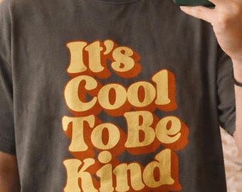 Könnte beinhalten: Pfefferfarbenes T-Shirt mit dem Text "It's Cool To Be Kind" in einem Retro-Schriftzug. Der Text ist gelb und orange. Das Shirt ist dunkelgrau und hat eine lockere Passform.