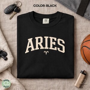 May include: Black t-shirt with the word "ARIES" in a vintage, off-white, collegiate-style font. The Aries zodiac symbol is below the word. The shirt is displayed on a neutral surface with keys, a basketball, and a lighter.