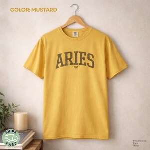 May include: Mustard-colored t-shirt with the word "ARIES" in a distressed, dark brown font. The Aries zodiac symbol is below the word. The shirt is hanging on a wooden hanger against a neutral background. The shirt is part of the Wholesome Tees Shop collection.