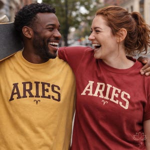 May include: Two people wearing matching Aries t-shirts. One shirt is mustard yellow, the other is red. The word "ARIES" is printed in a vintage style font across the front of each shirt, with the Aries symbol below.