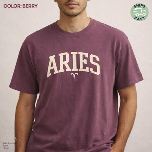 May include: Berry-colored t-shirt with the word "ARIES" in a white, collegiate-style font across the chest. The Aries zodiac symbol is below the word. The shirt has short sleeves and a crew neck.