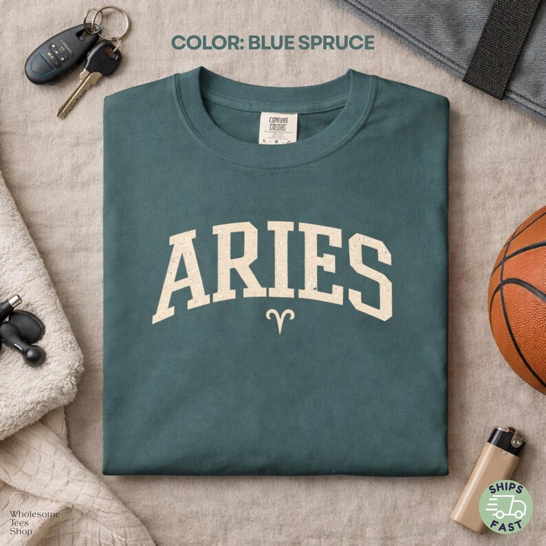 May include: Blue spruce-colored Aries t-shirt with a cream-colored Aries text and the Aries symbol. The shirt is folded and displayed with a basketball, keys, and a towel.