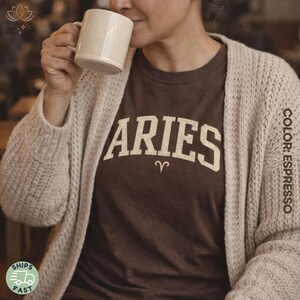 May include: A brown t-shirt with the word "ARIES" in a cream-colored, collegiate-style font, and the Aries zodiac symbol. The shirt is worn with a beige cardigan. A person is holding a cream-colored mug.