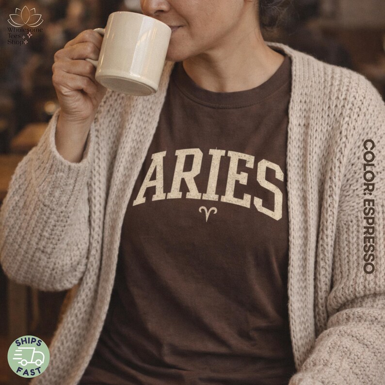May include: A brown t-shirt with the word "ARIES" in a cream-colored, collegiate-style font, and the Aries zodiac symbol. The shirt is worn with a beige cardigan. A person is holding a cream-colored mug.