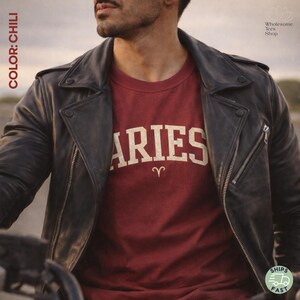 May include: A burgundy Aries t-shirt with a vintage design, paired with a black leather jacket. The shirt features the word "ARIES" in a bold, off-white font, with the Aries symbol below. The jacket has a classic biker style.