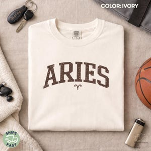 May include: Ivory-colored t-shirt with the word "ARIES" in a brown, distressed font. The Aries zodiac symbol is below the word. The shirt is folded on a neutral surface, with keys, a basketball, and other items nearby.