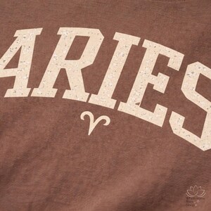 May include: Close-up of a brown fabric with the word "ARIES" in a distressed, off-white font. Below the word is the Aries zodiac symbol. The image has a vintage, textured look.