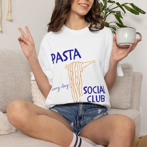 May include: White t-shirt with the words "PASTA SOCIAL CLUB" in blue and orange. The shirt features a graphic of pasta being held by a fork. The words "every day" are written below the word "PASTA".