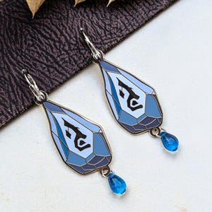 Peut inclure: Une paire de boucles d'oreilles pendantes argentées avec une breloque en forme de goutte ornée d'un motif géométrique bleu et blanc à facettes. Chaque breloque présente un symbole noir stylisé au centre et pend à un anneau argenté avec une perle bleue.