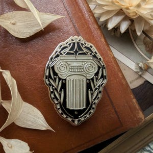 Peut inclure: Épingle ovale avec une bordure argentée et un intérieur noir. L'épingle présente un motif de colonne stylisée et détaillée en argent, avec un sommet décoratif et une tige cannelée. La bordure comprend un motif de feuilles et géométrique.