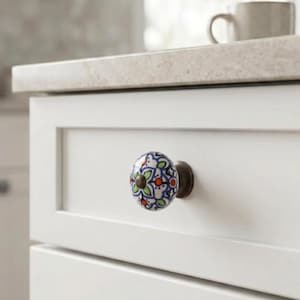 May include: A round ceramic drawer knob featuring a hand-painted floral pattern in blue, green, and orange on a white background, attached to a dark metal base and mounted on a white cabinet drawer front with a recessed panel design.