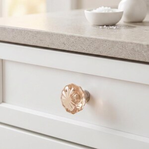 May include: A round, faceted, light pink glass cabinet knob with a metallic base, installed on a white drawer front. The knob features a radial, star-like pattern cut into the glass, reflecting light with a soft, translucent, and polished appearance.