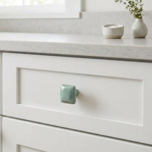 May include: A square cabinet knob with a speckled light green glaze and rounded edges, mounted on a white shaker-style drawer front. The knob features a glossy finish with subtle variations in color, providing a tactile grip for opening.