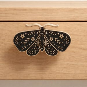 May include: A decorative black and silver moth-shaped drawer pull. The moth's wings are detailed with floral and arrow patterns. The drawer pull is mounted on a light-colored wooden drawer.