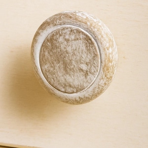 May include: A round, distressed cabinet knob with a light brown and white finish. The knob has a circular design with a recessed center and a white rim. The knob is made of wood and has a rustic appearance.