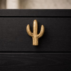 May include: A gold-colored cactus-shaped cabinet pull is mounted on a black wooden drawer. The cabinet pull is a detailed, three-dimensional design, adding a decorative touch to the furniture. The black wood grain is visible.