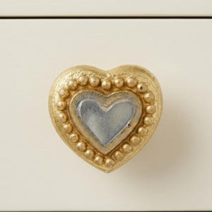 May include: A gold-colored heart-shaped cabinet knob with a silver-colored heart in the center. The knob has a beaded border and is set against a cream-colored background. Decorative hardware.