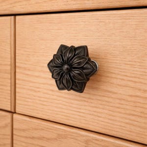 May include: A dark bronze flower-shaped cabinet knob. The knob is mounted on a light-colored wooden cabinet. The flower design features detailed petals and a central button. This decorative hardware adds a touch of elegance to furniture.