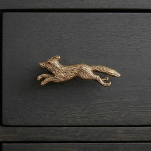 May include: A bronze-colored fox-shaped drawer pull in a running pose. The fox is detailed with textured fur and is mounted on a dark gray wooden drawer front. The hardware adds a rustic, animal-themed accent.