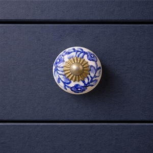 May include: A round, white ceramic cabinet knob with a blue floral design and a gold-colored metal center. The knob is mounted on a dark blue cabinet door, showcasing a classic decorative element.